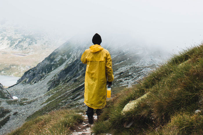 A hiker wearing a bright yellow raincoat and backpack walking along a steep mountain ridge under cloudy skies.